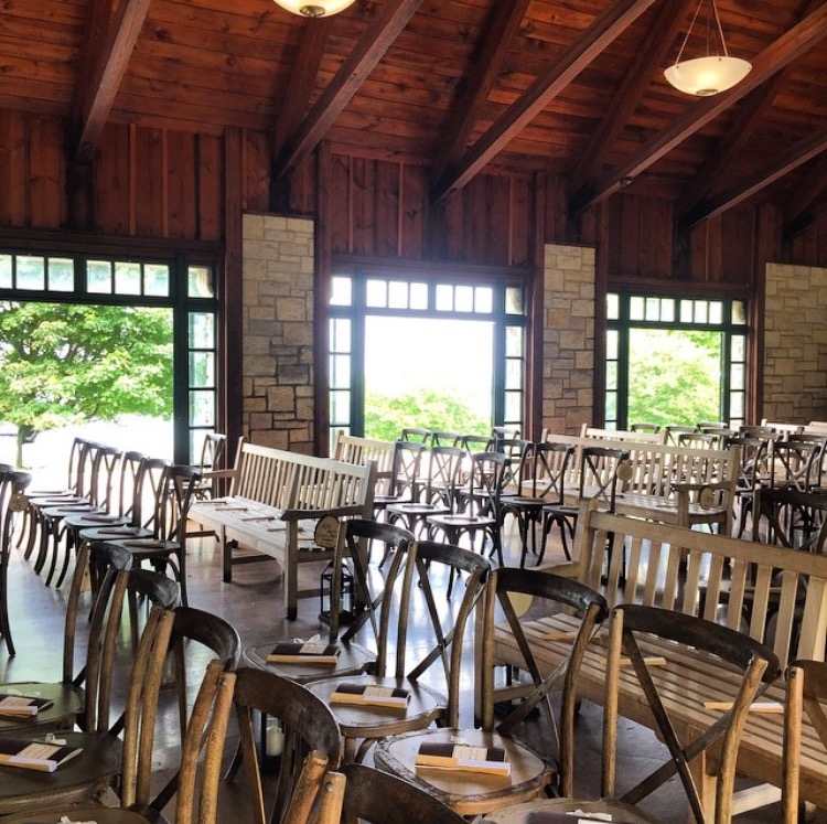 Wooden benches and chairs arranged in rows in a lodge with large windows and trees outside.